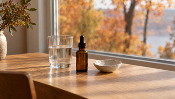 Eikenhouten tafel bij het raam in herfstochtendlicht met een glas water, een merkloos amberkleurig pipetflesje en een keramisch schaaltje, minoxidil tabletten of lotion